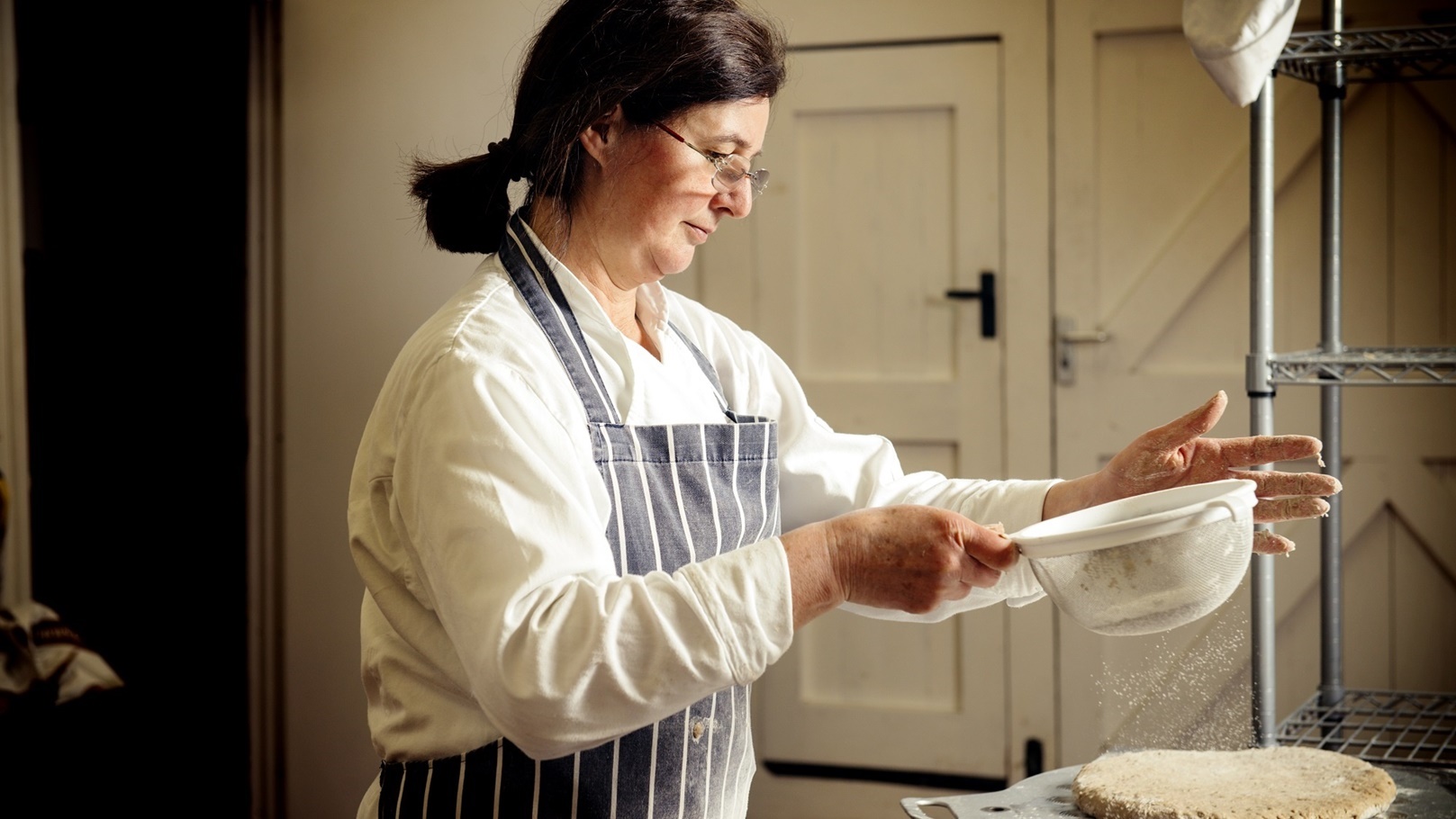 female-baker-sprinkling-flour-on-bread-dough-at-ba-2022-03-09-02-11-44-utc